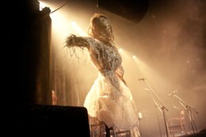 Postać w białym kostiumie z traw i płótna podczas koncertu Jare Gody na festiwalu Folkowe Ucho, oświetlona ciepłym światłem reflektorów.